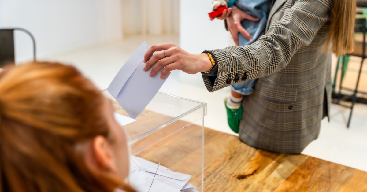 A person holding a child with their right hand and placing a white paper voting ballot into a box with their left hand.